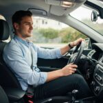 Mladý muž sedí ve voze připraven k jízdě, ruce na volantu, klidný výraz, denní světlo v interiéru auta.