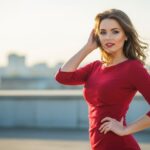 Mladá žena v červených šatech s úsměvem, venku s rozmazaným městským pozadím.