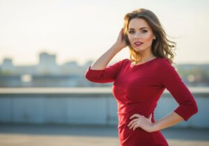 Mladá žena v červených šatech s úsměvem, venku s rozmazaným městským pozadím.
