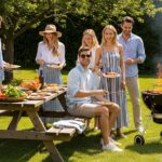 Skupina přátel na grilování na zahradě v letním oblečení u stolu s jídlem a grilem ve slunečném dni