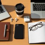 Moderní pracovní stůl s notebookem, telefonem, kávou, poznámkovým blokem, náušnicemi a brýlemi z vrchu